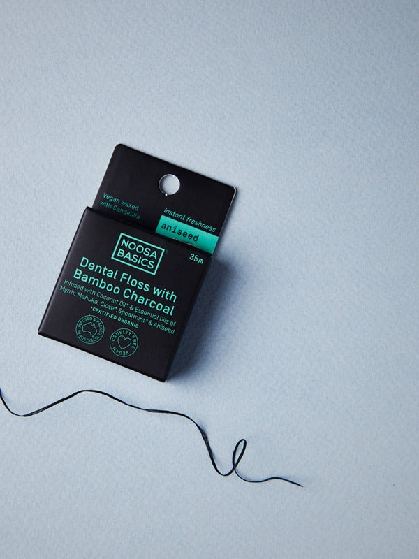 Black Noosa Basics Aniseed Bamboo Charcoal Dental Floss carton on a pale blue surface with a loose length of dark floss curling beside it.