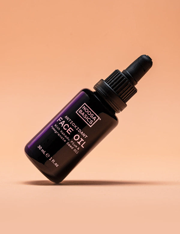 Dark Noosa Basics Antioxidant Face Oil bottle with a black dropper cap tilted diagonally against a peach background, casting a small shadow beneath it.