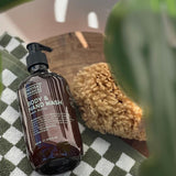Amber Noosa Basics Lemon Myrtle Body and Hand Wash pump bottle lying on a green-and-white checked cloth beside a natural sponge, framed by large green leaves.