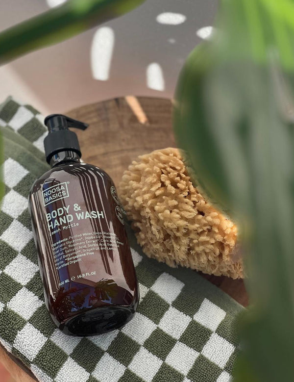 Amber Noosa Basics Lemon Myrtle Body and Hand Wash pump bottle lying on a green-and-white checked cloth beside a natural sponge, framed by large green leaves.