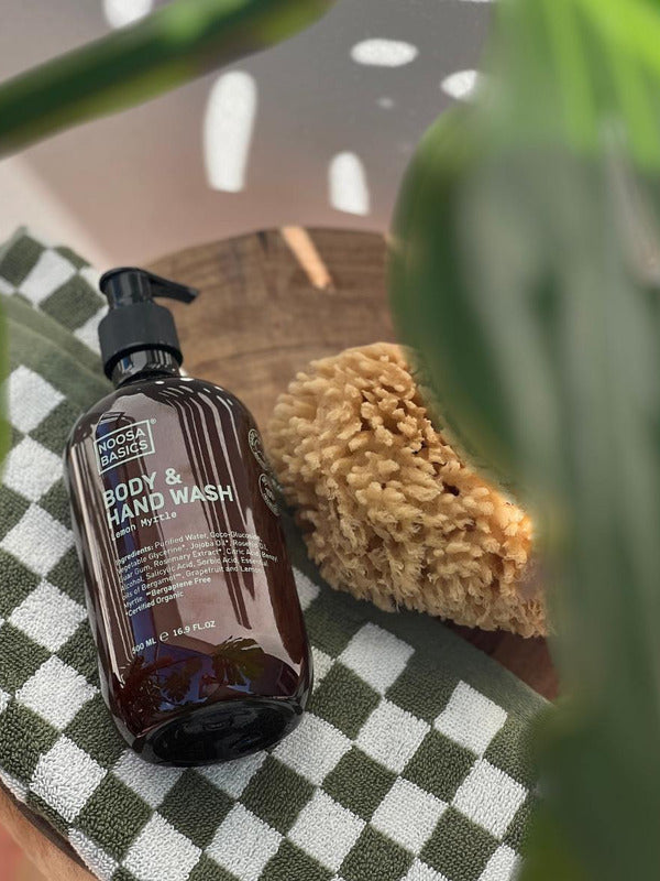 Amber Noosa Basics Lemon Myrtle Body and Hand Wash pump bottle lying on a green-and-white checked cloth beside a natural sponge, framed by large green leaves.