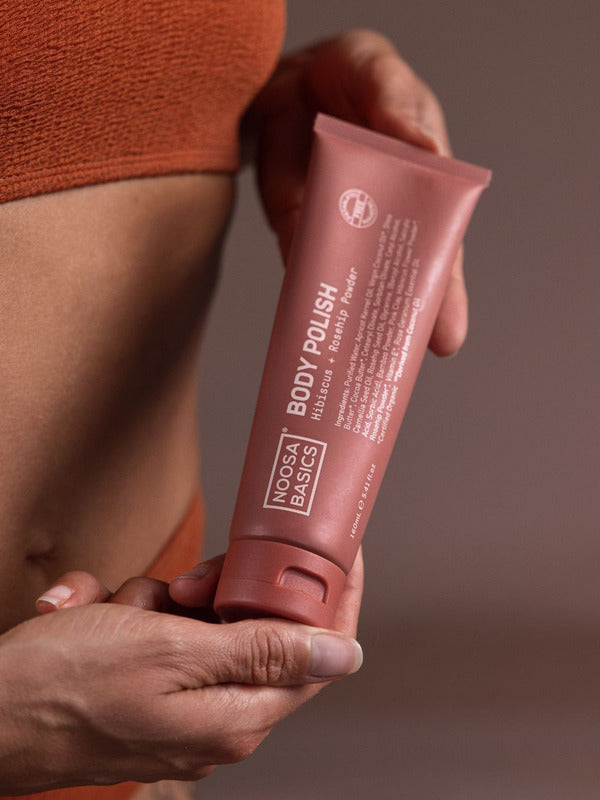 Person holding a dusty pink Noosa Basics Hibiscus and Rosehip Body Polish tube in both hands against their bare abdomen below a rust-coloured top.