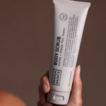 Hand holding a tall beige Noosa Basics Coffee and Orange Peel Body Scrub tube upright, with black lettering running along the front.