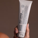 Hand holding a tall beige Noosa Basics Coffee and Orange Peel Body Scrub tube upright, with black lettering running along the front.