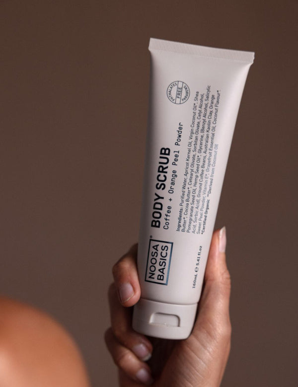 Hand holding a tall beige Noosa Basics Coffee and Orange Peel Body Scrub tube upright, with black lettering running along the front.