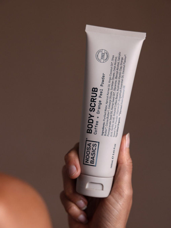 Hand holding a tall beige Noosa Basics Coffee and Orange Peel Body Scrub tube upright, with black lettering running along the front.