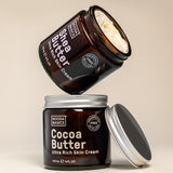 Open Noosa Basics Cocoa Butter and Shea Butter skin cream jars stacked with their silver lids on a warm beige surface.