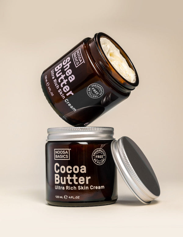 Open Noosa Basics Cocoa Butter and Shea Butter skin cream jars stacked with their silver lids on a warm beige surface.