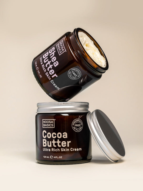 Open Noosa Basics Cocoa Butter and Shea Butter skin cream jars stacked with their silver lids on a warm beige surface.