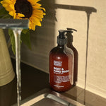 Amber Noosa Basics Coconut and Lime Body and Hand Wash pump bottle beside a kitchen sink and a vase of sunflowers in warm sunlight.