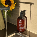 Amber Noosa Basics Coconut and Lime Body and Hand Wash pump bottle beside a kitchen sink and a vase of sunflowers in warm sunlight.