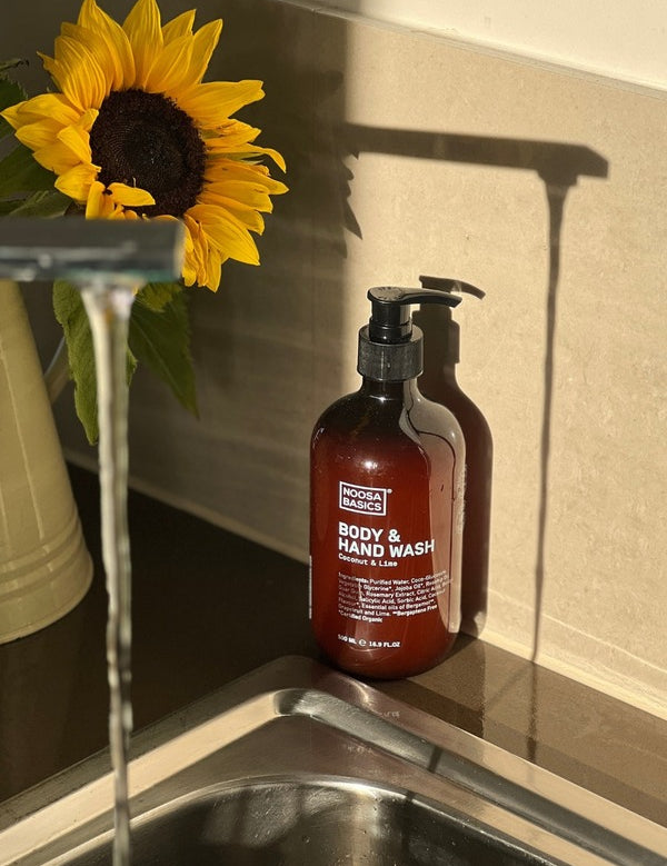 Amber Noosa Basics Coconut and Lime Body and Hand Wash pump bottle beside a kitchen sink and a vase of sunflowers in warm sunlight.