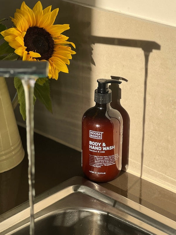 Amber Noosa Basics Coconut and Lime Body and Hand Wash pump bottle beside a kitchen sink and a vase of sunflowers in warm sunlight.