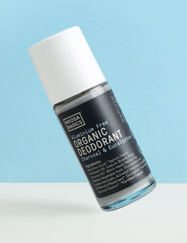 Clear Noosa Basics Charcoal and Eucalyptus roll-on deodorant bottle with a black label and white cap tilted diagonally against a pale blue background.