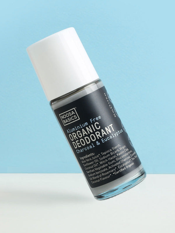 Clear Noosa Basics Charcoal and Eucalyptus roll-on deodorant bottle with a black label and white cap tilted diagonally against a pale blue background.