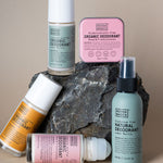 Five Noosa Basics deodorants arranged around a dark rock: three roll-on bottles with green, orange and pink labels, a square pink tin, and a pale green spray bottle.