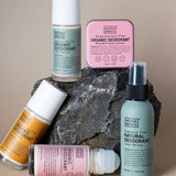 Five Noosa Basics deodorants arranged around a dark rock: three roll-on bottles with green, orange and pink labels, a square pink tin, and a pale green spray bottle.
