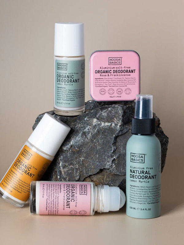 Five Noosa Basics deodorants arranged around a dark rock: three roll-on bottles with green, orange and pink labels, a square pink tin, and a pale green spray bottle.