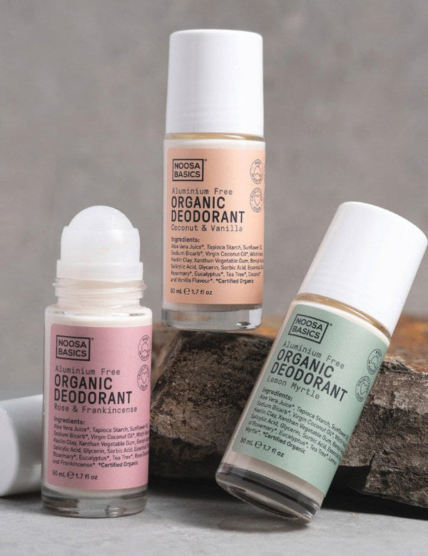 Three Noosa Basics roll-on deodorants arranged around a rock: peach-labelled Coconut and Vanilla, green-labelled Lemon Myrtle, and pink-labelled Rose and Frankincense with its roller exposed and cap beside it.