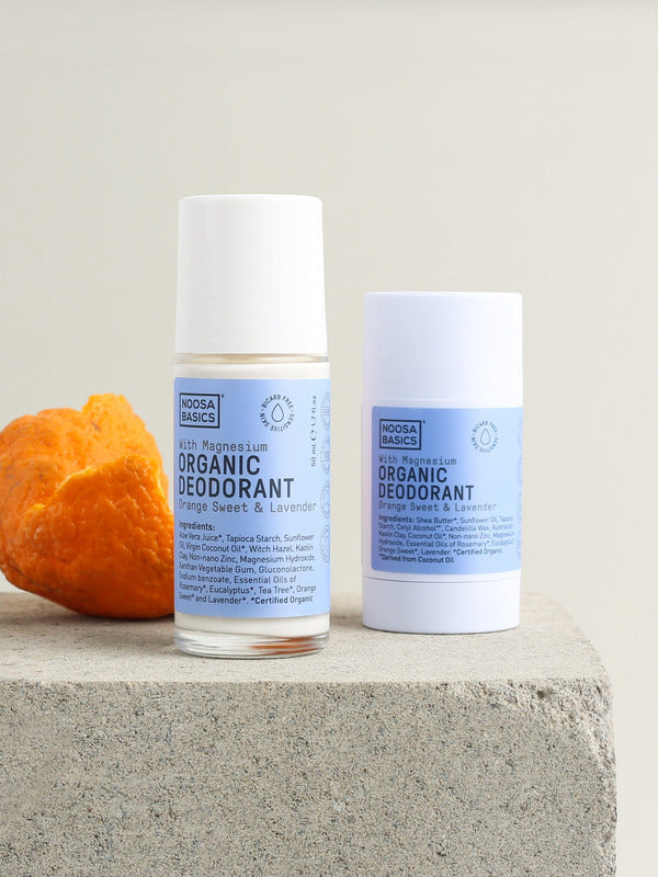 Noosa Basics Sweet Orange and Lavender roll-on and stick deodorants with pale blue labels and white caps, standing on a square stone block beside curled orange peel.