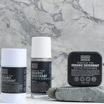 Noosa Basics Charcoal and Eucalyptus deodorant stick, roll-on bottle and square tin arranged beside a flat grey stone against a veined marble background, all with black labels.