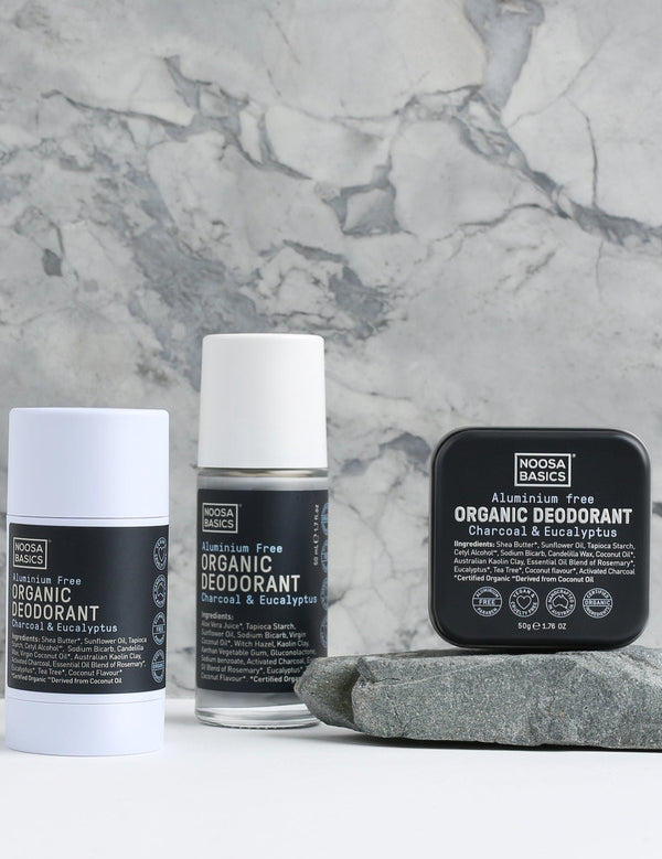 Noosa Basics Charcoal and Eucalyptus deodorant stick, roll-on bottle and square tin arranged beside a flat grey stone against a veined marble background, all with black labels.