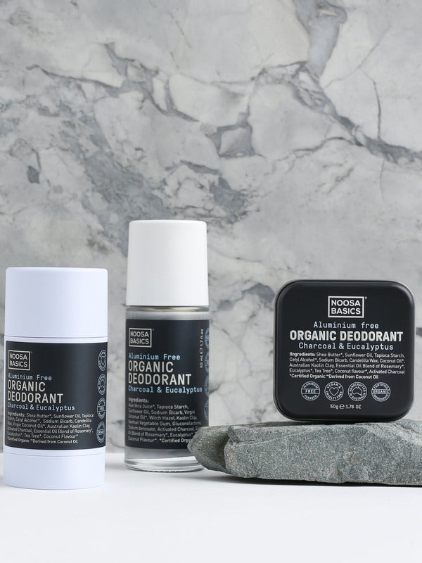 Noosa Basics Charcoal and Eucalyptus deodorant stick, roll-on bottle and square tin arranged beside a flat grey stone against a veined marble background, all with black labels.