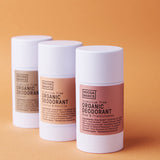 Three white Noosa Basics deodorant sticks standing in a staggered row against orange, with beige, peach and pink labels; Coconut and Vanilla and Rose and Frankincense are visible on the front two.