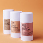Three white Noosa Basics deodorant sticks standing in a staggered row against orange, with beige, peach and pink labels; Coconut and Vanilla and Rose and Frankincense are visible on the front two.