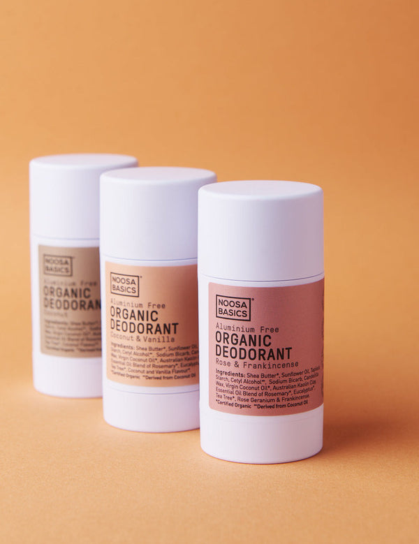 Three white Noosa Basics deodorant sticks standing in a staggered row against orange, with beige, peach and pink labels; Coconut and Vanilla and Rose and Frankincense are visible on the front two.