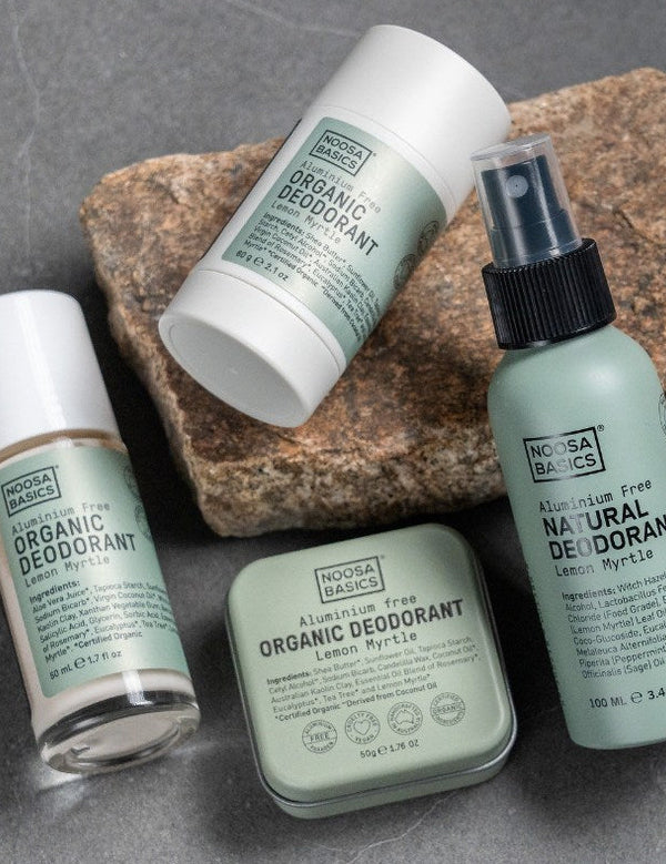 Noosa Basics Lemon Myrtle deodorant stick, roll-on bottle, spray and tin arranged across layered natural stones with the green-labelled stick resting diagonally in front.