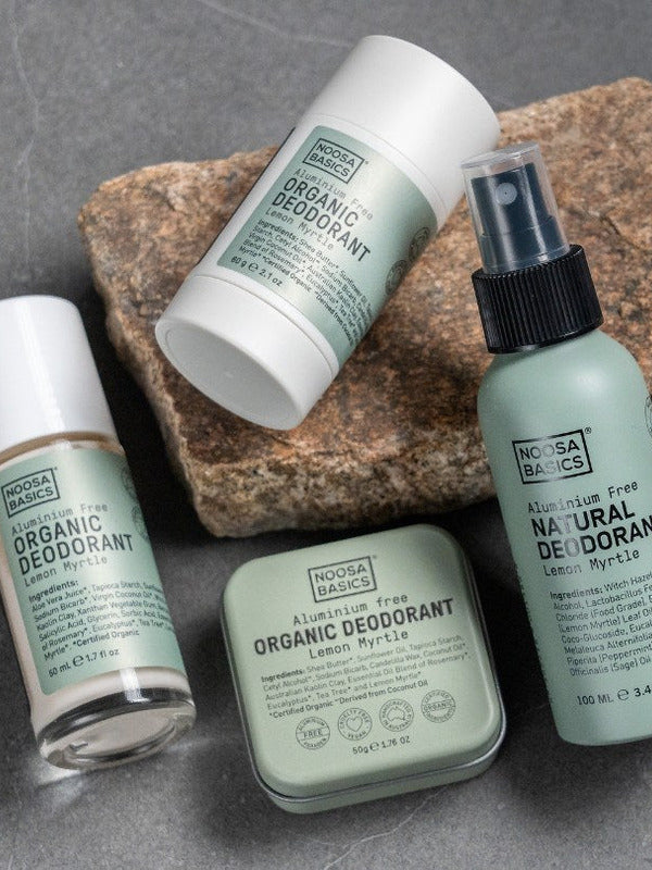 Noosa Basics Lemon Myrtle deodorant stick, roll-on bottle, spray and tin arranged across layered natural stones with the green-labelled stick resting diagonally in front.