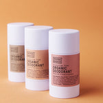 Three white Noosa Basics natural deodorant sticks, including Sandalwood, arranged in a staggered group against a warm orange background.