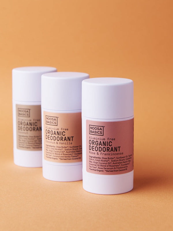 Three white Noosa Basics natural deodorant sticks, including Sandalwood, arranged in a staggered group against a warm orange background.