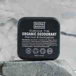 Closed black square Noosa Basics Charcoal and Eucalyptus deodorant tin standing upright on a flat grey stone against a veined marble background.