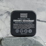 Closed black square Noosa Basics Charcoal and Eucalyptus deodorant tin standing upright on a flat grey stone against a veined marble background.