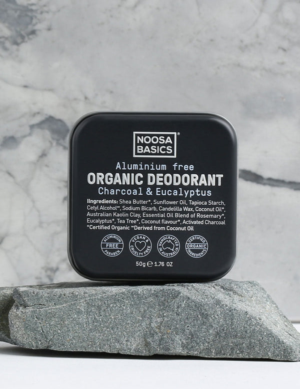 Closed black square Noosa Basics Charcoal and Eucalyptus deodorant tin standing upright on a flat grey stone against a veined marble background.