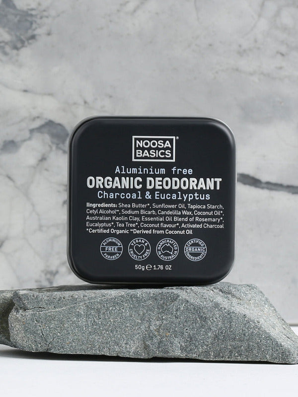 Closed black square Noosa Basics Charcoal and Eucalyptus deodorant tin standing upright on a flat grey stone against a veined marble background.