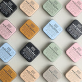 Sixteen square Noosa Basics deodorant tins arranged diagonally in a four-by-four grid, with black, peach, green, pink, orange, blue and beige lids facing upwards.