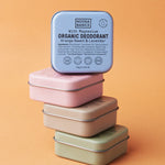 Four square Noosa Basics deodorant tins stacked against orange: green, peach and pink tins lie flat below an upright blue Sweet Orange and Lavender tin.