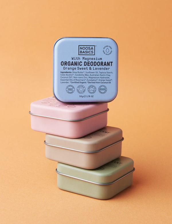 Four square Noosa Basics deodorant tins stacked against orange: green, peach and pink tins lie flat below an upright blue Sweet Orange and Lavender tin.