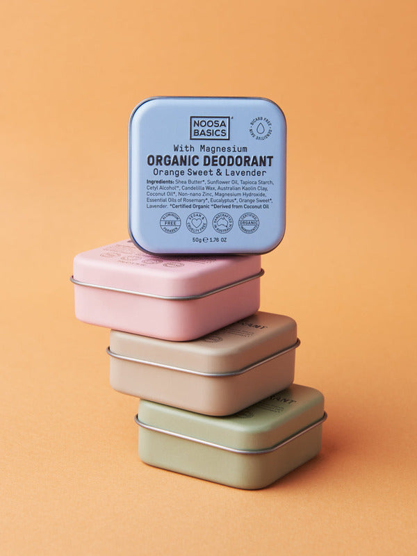 Four square Noosa Basics deodorant tins stacked against orange: green, peach and pink tins lie flat below an upright blue Sweet Orange and Lavender tin.