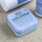 Close-up of pale blue Noosa Basics Sweet Orange and Lavender deodorants on a stone surface, with a square tin in front of a white stick container and a roll-on bottle.