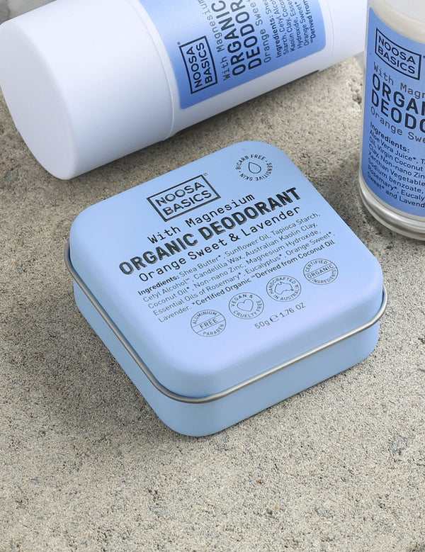 Close-up of pale blue Noosa Basics Sweet Orange and Lavender deodorants on a stone surface, with a square tin in front of a white stick container and a roll-on bottle.