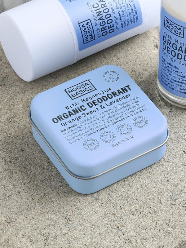 Close-up of pale blue Noosa Basics Sweet Orange and Lavender deodorants on a stone surface, with a square tin in front of a white stick container and a roll-on bottle.
