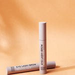 Two slim beige Noosa Basics Eye Lash and Brow Revitaliser tubes arranged on a warm orange background with long shadows.