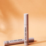 Two slim beige Noosa Basics Eye Lash and Brow Revitaliser tubes arranged on a warm orange background with long shadows.
