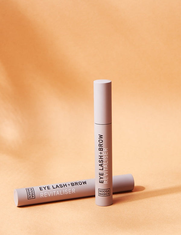 Two slim beige Noosa Basics Eye Lash and Brow Revitaliser tubes arranged on a warm orange background with long shadows.