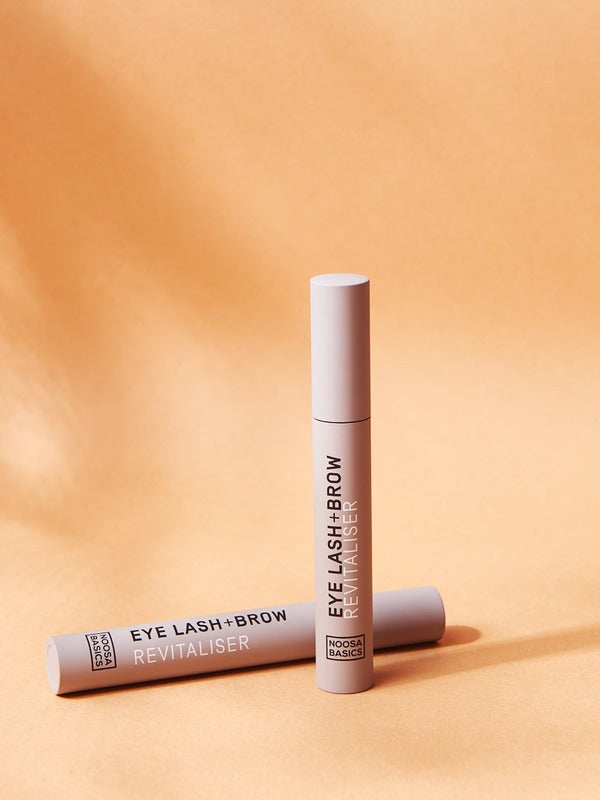 Two slim beige Noosa Basics Eye Lash and Brow Revitaliser tubes arranged on a warm orange background with long shadows.