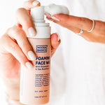 Hands holding Noosa Basics Foaming Face Wash and dispensing a mound of white cleansing foam onto the fingertips.